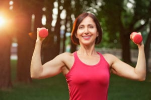 Portrait of cheerful aged woman in fitness wear exercising with red dumbbells in park.